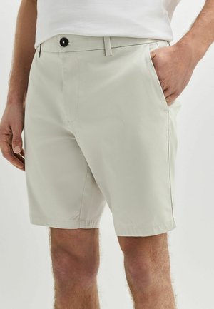 Férfi világos bézs színű térdig érő rövidnadrágot visel, gombbal és cipzárral, jobb zsebében a keze, fehér ing betűrve.