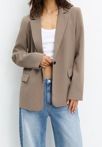 Woman wearing loose beige blazer, white cropped tank top, and light blue high-waisted jeans against plain background.