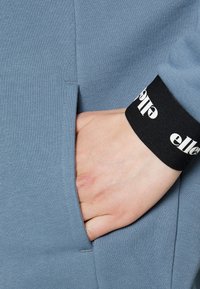 Blå, ribbad tyg med en sidoficka; svart mudd med vit text. Närbild av en hand som vilar i fickan.