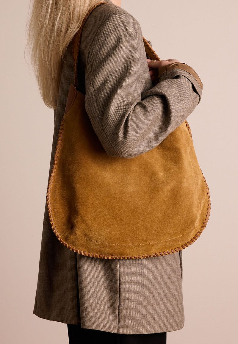 Suede shoulder bag in mustard yellow with a soft texture, rounded shape, and contrasting stitched edges; paired with a gray blazer.
