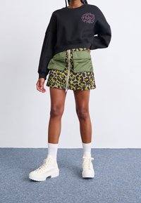 Sweatshirt noir avec logo violet, jupe imprimée léopard verte avec fermeture éclair, et bottes blanches montantes. Couches texturées et poches surdimensionnées.