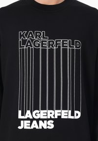 Svart hettegenser med kraftig hvit tekst som leser "KARL LAGERFELD" og "LAGERFELD JEANS," med vertikale linjeaksenter gjennomgående.