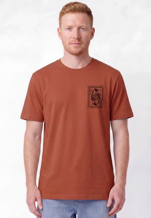 Mann mit heller Haut und kurzen roten Haaren trägt ein rostfarbenes T-Shirt mit einem schwarzen Joker-Spielkarten-Design auf der linken Brust.
