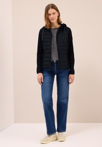 Vadderad svart jacka med huva och dragkedja, i kombination med en randig långärmad tröja, blå jeans och ljusa sneakers.