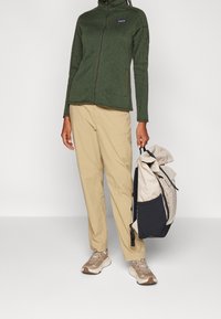 Grön fleecejacka med dragkedja framtill, beige byxor och lättviktig ryggsäck. Sneakers är bruna med inslag av vitt och bruna accenter.