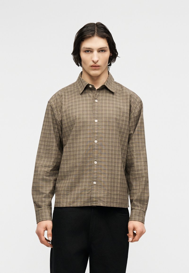 Νεαρό άτομο με μεσαίου μήκους σκούρα μαλλιά φορώντας ένα καφέ καρό πουκάμισο με μακριά μανίκια και μαύρο παντελόνι, στέκεται μπροστά σε απλό φόντο.