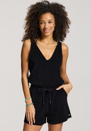 Keharuldse juustega naine, kes kannab musta v-kaelusega varrukateta kombinesooni, millel on nööriga lühikesed püksid, naeratades ja poseerides ühe käega taskus.