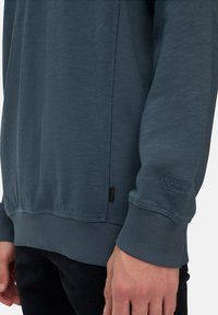 Jasnoniebieski sweter z ribbowymi mankietami, teksturowana tkanina i naszywka z logiem na rękawie. Posiada prosty, sportowy design.