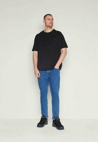 Czarna koszulka krótki rękaw, niebieskie dżinsy, czarne sneakersy. Model stoi prosto na neutralnym tle, z rękami w kieszeniach.