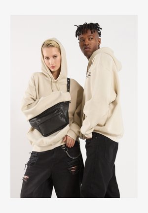 Felpe beige con ampi tasconi a marsupio, abbinate a jeans neri strappati. Un modello tiene una borsa a tracolla in pelle nera.