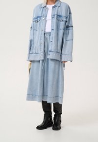 Vaaleansininen ylisuurikokoinen farkkutakki ja siihen sopiva hame yhdistettynä mustiin keskipitkiin saappaisiin ja valkoiseen puseroon, pitäen kädessä pientä beigeä laukkua.