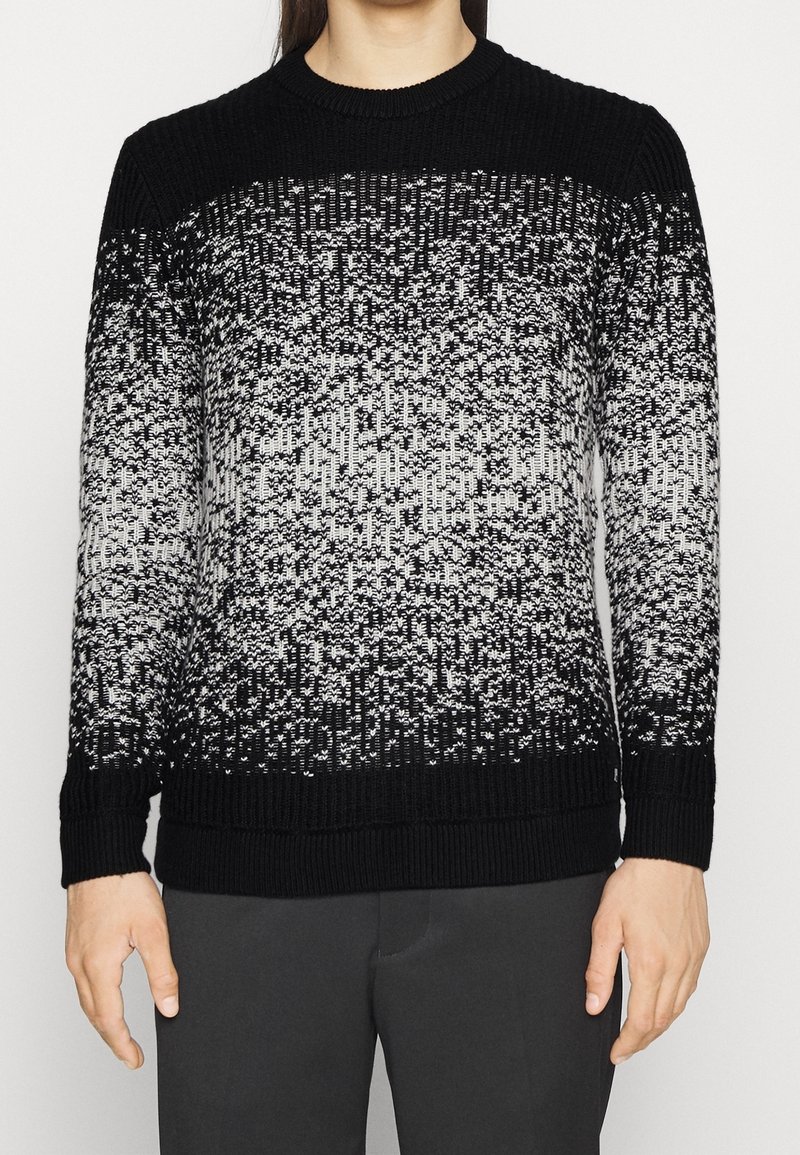 Homme portant un pull en maille dégradé noir et blanc à manches longues et un pantalon noir, debout devant un fond clair uni.