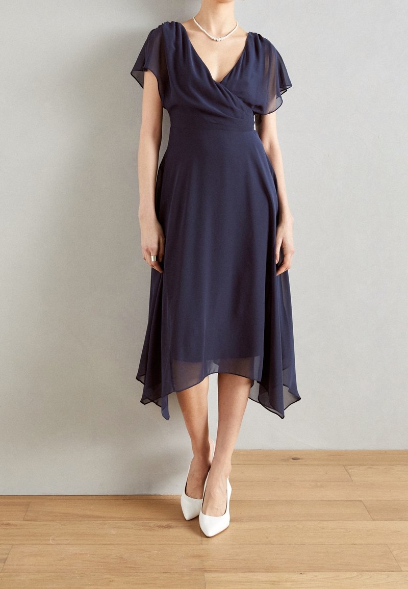 Marinblå chiffonomslagsklänning med korta volangärmar, v-ringning och asymmetrisk nederdel. Kombinerad med vita spetsiga klackskor.