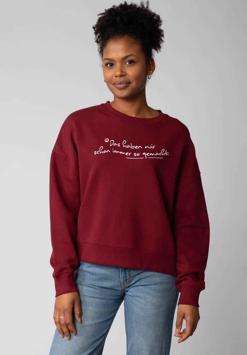 Vrouw met krullend haar die een bordeauxrode sweatshirt draagt met de Duitse tekst "Das haben wir schon immer so gemacht" en lichtblauwe jeans.