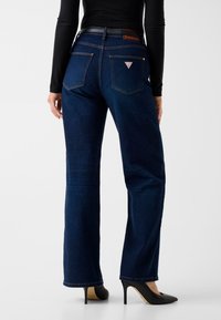 Jean flare en denim bleu foncé à taille haute, avec un patch logo au dos, souligné par des coutures dorées et une ceinture noire.