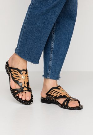 Sandales en cuir noir et tan avec un design découpé en forme d'ailes, accentuées par un détail en cœur doré et une semelle noire cloutée, portées avec un jean effiloché.
