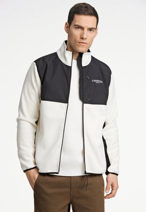 Hvid og sort zip-up jakke med høj krave, der har en glat tekstur, lodrette syninger og logo på brystet, parret med en strikket sweater.