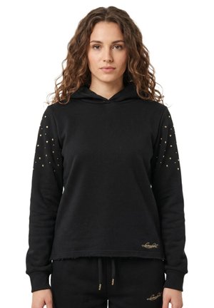 Une femme présente un sweat-shirt noir à capuche orné de clous dorés sur les manches, avec un pantalon noir assorti, sur fond blanc.
