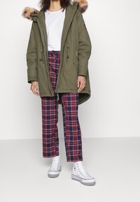 Parka verde oliva con cappuccio bordato di pelliccia, abbinata a pantaloni a quadri blu navy e rossi e sneakers alte bianche. Coulisse regolabili, chiusura con zip.