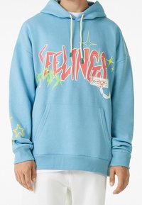 Sudadera azul clara con cordones blancos, texto "FEELINGS" en rojo y amarillo, gráficos abstractos y estrellas amarillas en las mangas.