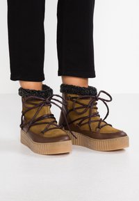 Botas cortas en marrón y marrón claro con suelas de goma texturizadas y un puño negro de felpa. Presenta cordones gruesos y un diseño de plataforma.