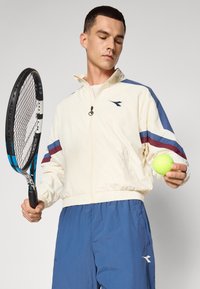 Gaiši krēmkrāsas zip-up jaka ar ziliem un sarkaniem svītrām, pārī ar zilām sporta šortiem; tur rokās zaļu tenisa bumbu un raketi.