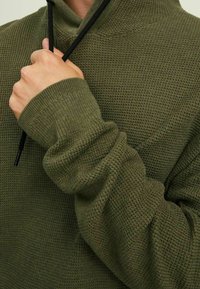 Zielony, teksturowany sweter z wysokim kołnierzem. Ma detal ze sznurkiem i ściągaczami przy mankietach. Zbliżenie na rękę trzymającą sznurek.