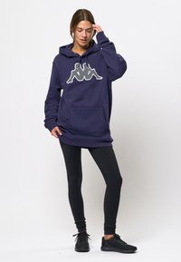 Sudadera navy con un gran logotipo blanco, presenta un bolsillo frontal y capucha con cordón, combinada con leggings negros y zapatillas deportivas negras.
