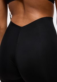 Κοντινό πλάνο της μέσης και των γοφών ενός ατόμου που φοράει μαύρο, χωρίς ραφές, ψηλόμεσο κολάν με ζώνη σε σχήμα V.