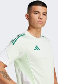 Maglietta sportiva verde chiaro con accenti bianchi e strisce verdi; presenta un collo rotondo e un piccolo logo sul petto. Texture liscia, maniche corte.