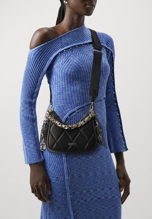 Femme portant une robe bleue côtelée à épaules dénudées avec manches longues et un sac bandoulière matelassé noir avec chaîne dorée et détails de foulard fleuri.
