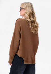 Bruine gebreide trui met een losse pasvorm, geribbelde onderkant en een ongelijkmatige onderste rand. Gecombineerd met donkere spijkerbroeken. Subtiele textuurdetail in de stof.