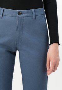 Ljust blå bomullsbyxor med knappstängning fram, sidofickor och smal passform. Sömmar synliga längs benen.
