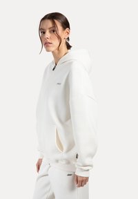 Hvid zip-up hættetrøje lavet af blød stof, med en stor hætte, forlommer og en subtil logo detalje. Vist med matchende bukser.