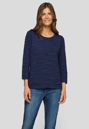 Lächelnde Frau in einem navy blauen, strukturierten Langarmoberteil und blauen Jeans, die vor einem einfarbigen hellgrauen Hintergrund steht.