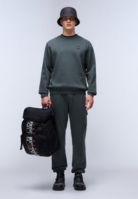 Donkergroen sweatshirt en bijpassende joggingbroek, gecombineerd met een zwarte bucket hat. Houdt een zwarte rugzak vast met grafische tekst en ritsen.