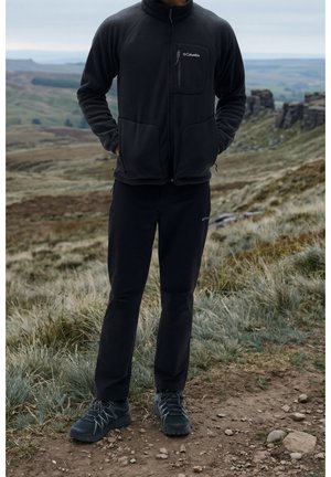 Persona con chaqueta negra Columbia, pantalones negros y zapatos de senderismo, de pie sobre terreno rocoso y con hierba, con colinas al fondo.