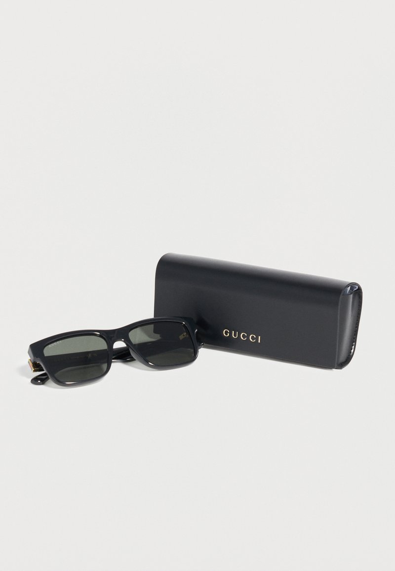 Black rectangular sunglasses with dark lenses beside a black Gucci case with gold lettering on a white background.