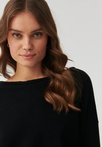 Maglione nero con scollo ampio e maniche morbide. Texture liscia e design rilassato, indossato da una modella con capelli mossi.