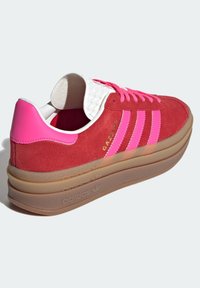 Czerwony zamszowy sneaker z różowymi akcentami i trzema różowymi paskami. Posiada biały teksturowany język oraz grubą, przejrzystą gumową podeszwę.