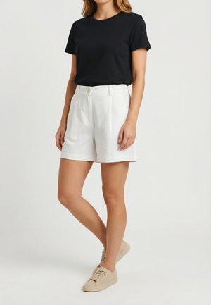 Mujer con camiseta negra de manga corta, pantalones cortos blancos de tiro alto con botones y zapatillas beige con cordones, de pie sobre fondo blanco.