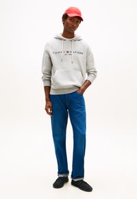 Sudadera gris con capucha y logo, pantalones vaqueros azules y zapatos negros. La sudadera tiene un bolsillo delantero y cordones. Una gorra roja completa el conjunto.