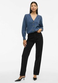 Modra dolg rokav cardigan z teksturiranimi bobble detajli, V-izrezom in gumbnim zapiranjem, v kombinaciji s črnimi hlačami visoke pasu in čevlji s peto.