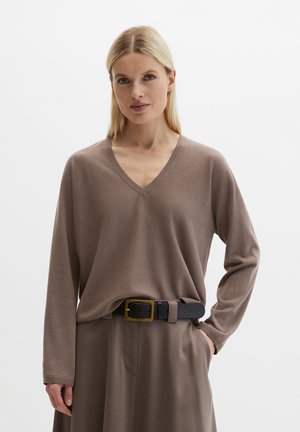 Frau mit glattem blondem Haar, die einen taupefarbenen V-Ausschnitt-Pullover trägt, der in passende, weit geschnittene Hosen mit einem schwarzen Gürtel gesteckt ist.