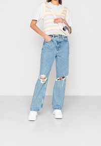 Gilet leggero a righe pastello sopra una maglietta bianca, abbinato a jeans destrutturati azzurri e sneakers bianche. Piccola borsa a tracolla.