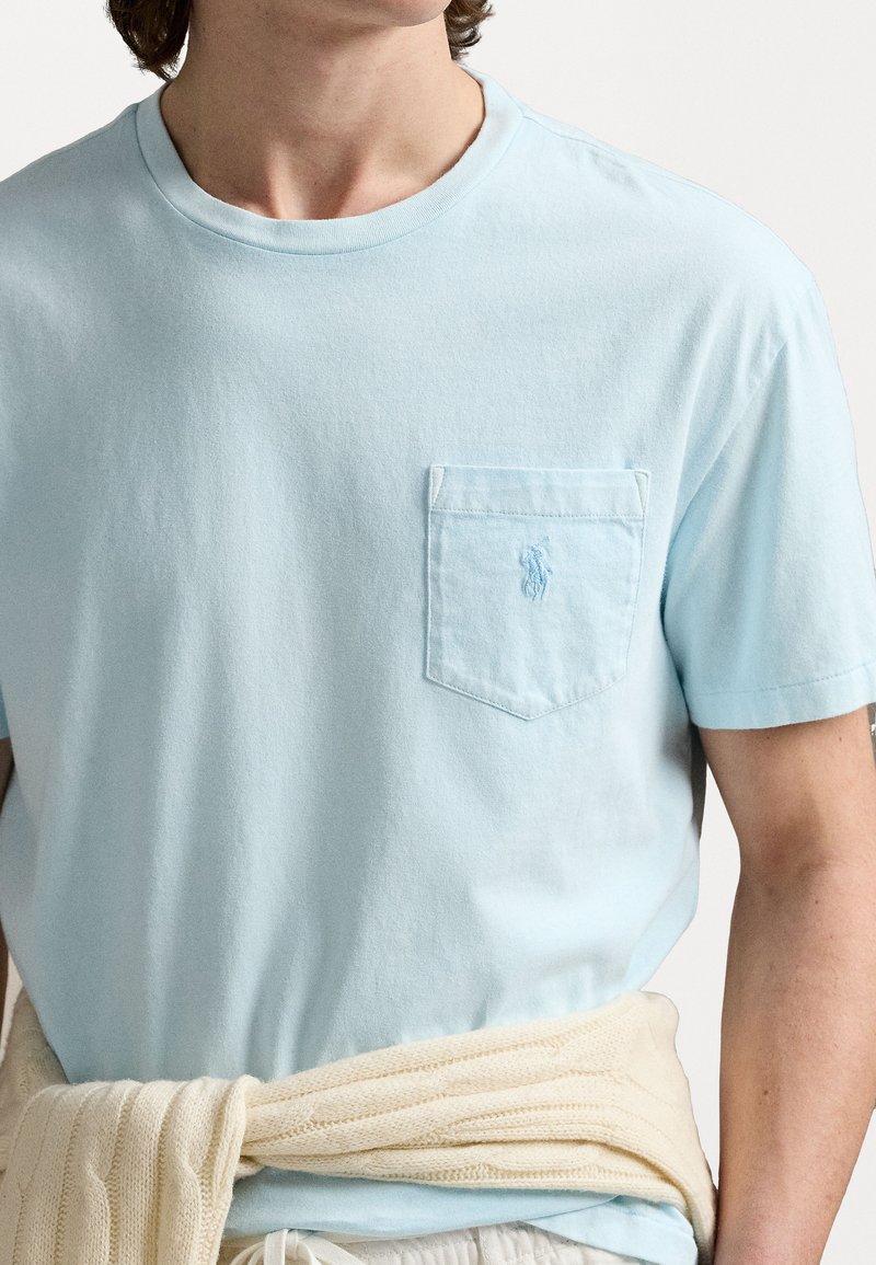 Jasnoniebieski bawełniany t-shirt z kieszonką na piersi, z wyszywanym logo. Okrągły dekolt, luzny krój, teksturowany materiał. Jasnobeżowy sweter związany w talii.