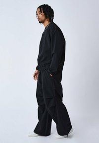 Schwarzer Sweatshirt und Hose, beide mit einer lockeren Passform. Der Stoff ist glatt mit elastischen Bündchen und Nähten. Kombiniert mit weißen Turnschuhen.