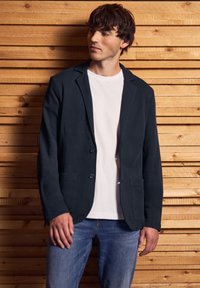 Marineblå blazer i texturerat tyg, med tvåknapprad och två framfickor, bärs över en vit t-shirt och matchas med blå jeans.