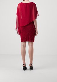 Czerwona sukienka koktajlowa z przezroczystą warstwą w formie peleryny, misternymi koronkowymi zdobieniami na spódnicy i falowanym dołem, zestawiona z czarnymi szpilkami.