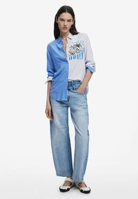 Camicia a righe blu e bianche con stampa grafica, abbinata a jeans a gamba larga blu chiaro e scarpe slip-on bianco e nere.
